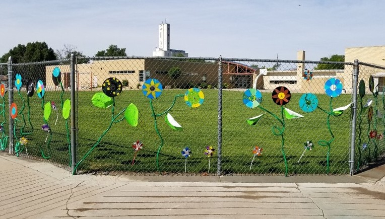 image of backside of a school with a lush green field, and chain link fence decorated with brightly colored flower sculptures