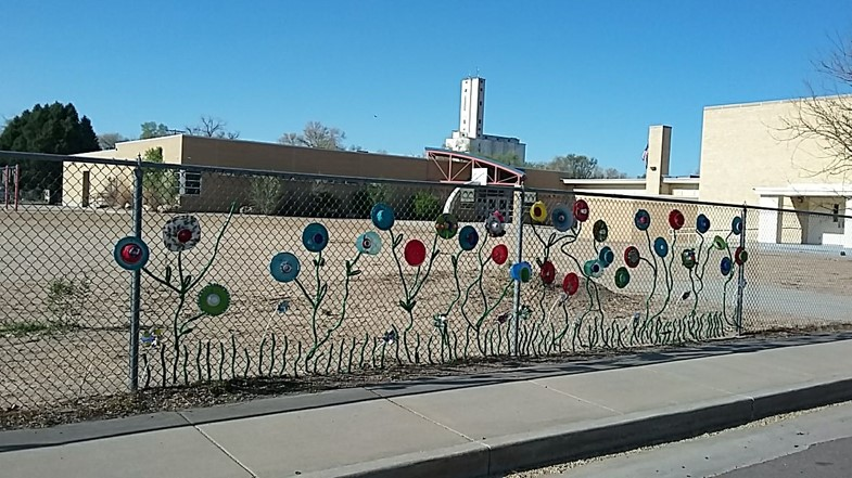 Brick school buildings with a dirt field surrounded by a chain link fence with flower sculptures as protest.